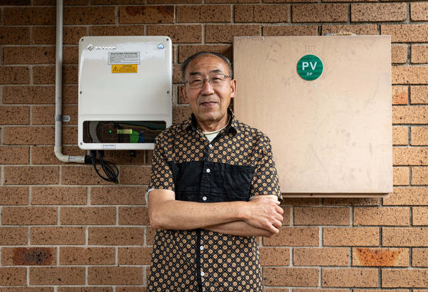 Elderly Asian man standing in front of his wall-mounted rooftop solar battery system.